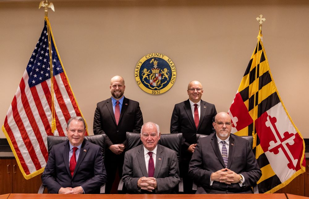 The current five Commissioners of St. Mary's County, seated and standing between the U.S. and Maryland flags in the Commissioner's meeting room.